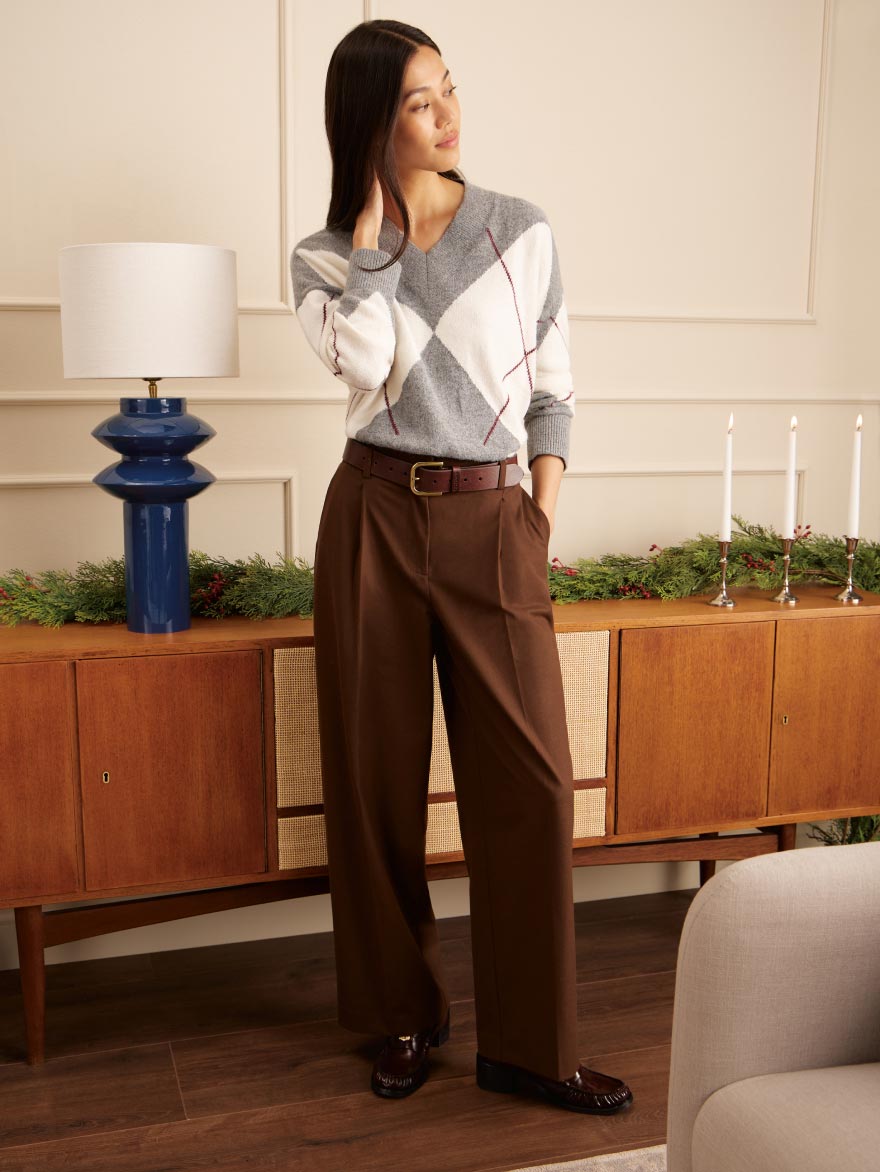 Model indoors beside holiday greenery wearing a grey and white argyle Tommy Hilfiger sweater with wide-leg brown trousers, leather belt, and loafers.