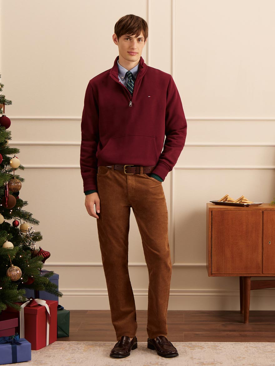 Model standing next to a decorated Christmas tree in a burgundy Tommy Hilfiger sweater over a blue shirt with brown trousers and loafers, gifts arranged at the base.