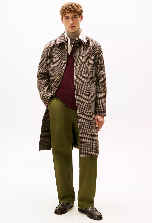 Full-body view of a model wearing a long patterned Tommy Hilfiger overcoat, styled over a burgundy sweater and green trousers.