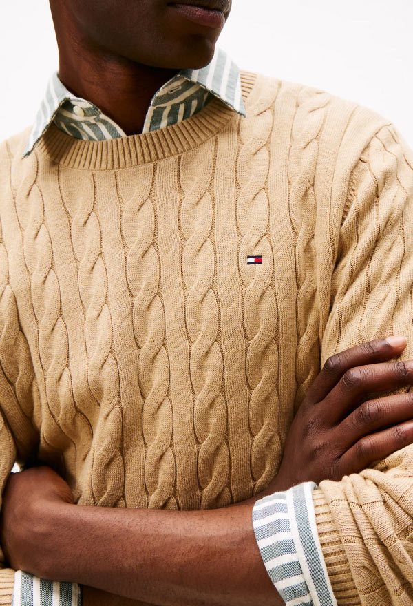 Close-up of a model wearing a beige cable-knit Tommy Hilfiger sweater layered over a green and white plaid shirt.