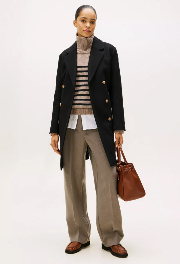 Model dressed in a navy double-breasted Tommy Hilfiger blazer over a striped top, paired with wide-leg trousers and holding a brown handbag.
