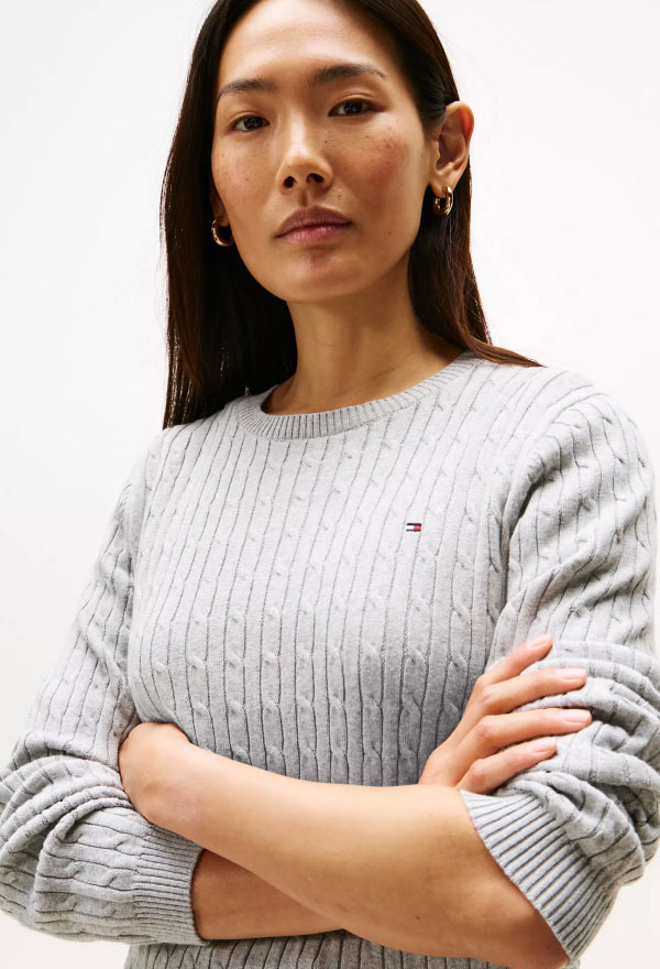 Model wearing a light gray cable-knit Tommy Hilfiger sweater with the embroidered flag logo on the chest.