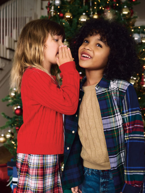 Two friends near a Christmas tree—one whispering to the other—wearing plaid and textured Tommy Hilfiger layers in a festive setting.