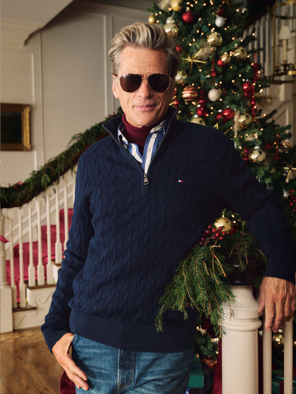 Man leaning on a banister in a navy cable-knit Tommy Hilfiger half-zip sweater and sunglasses beside a decorated tree.