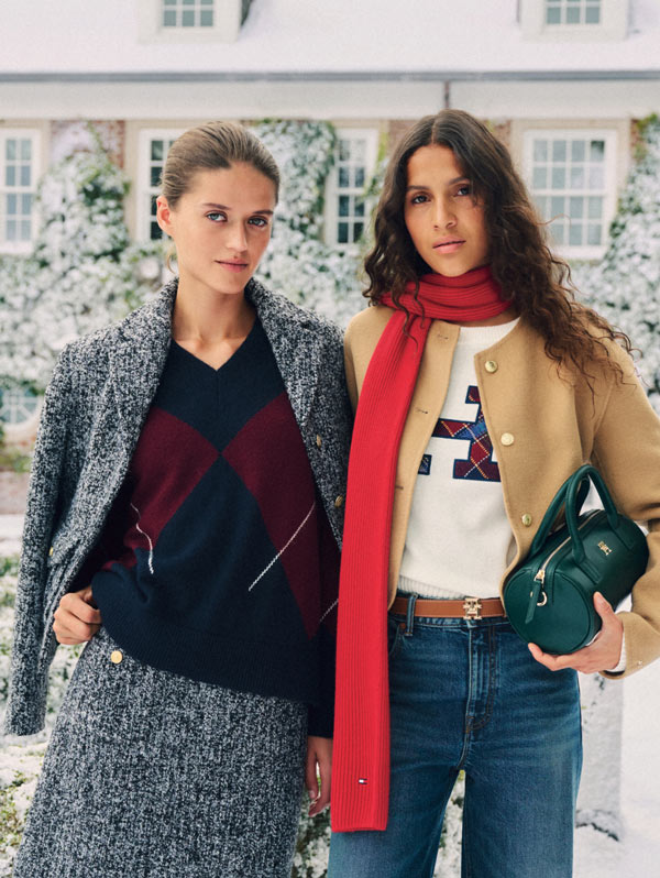 Two women on a winter street wearing Tommy Hilfiger layers—a gray coat with argyle sweater and red scarf, and a cream cardigan with red scarf and green handbag.