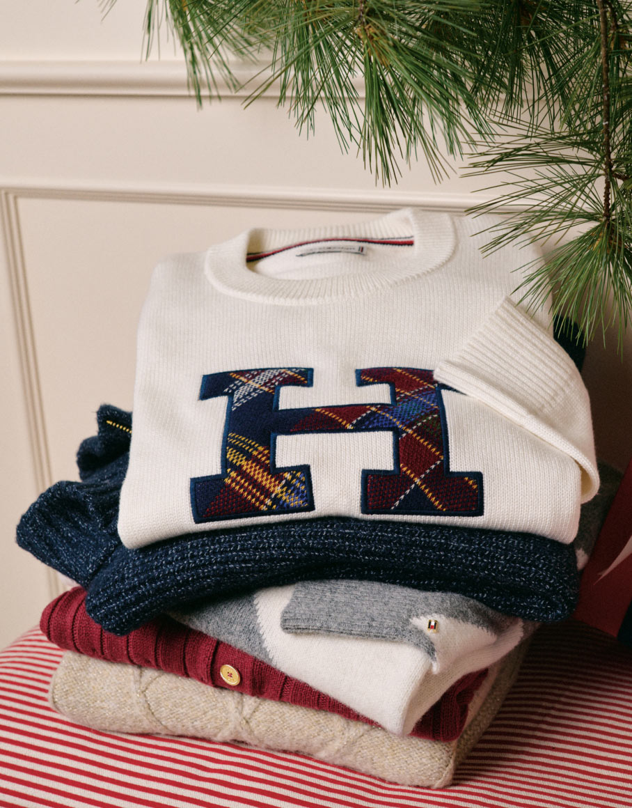Folded Tommy Hilfiger sweaters with varsity “H” logos stacked on a striped table beside ribbon and greenery in a holiday setting.