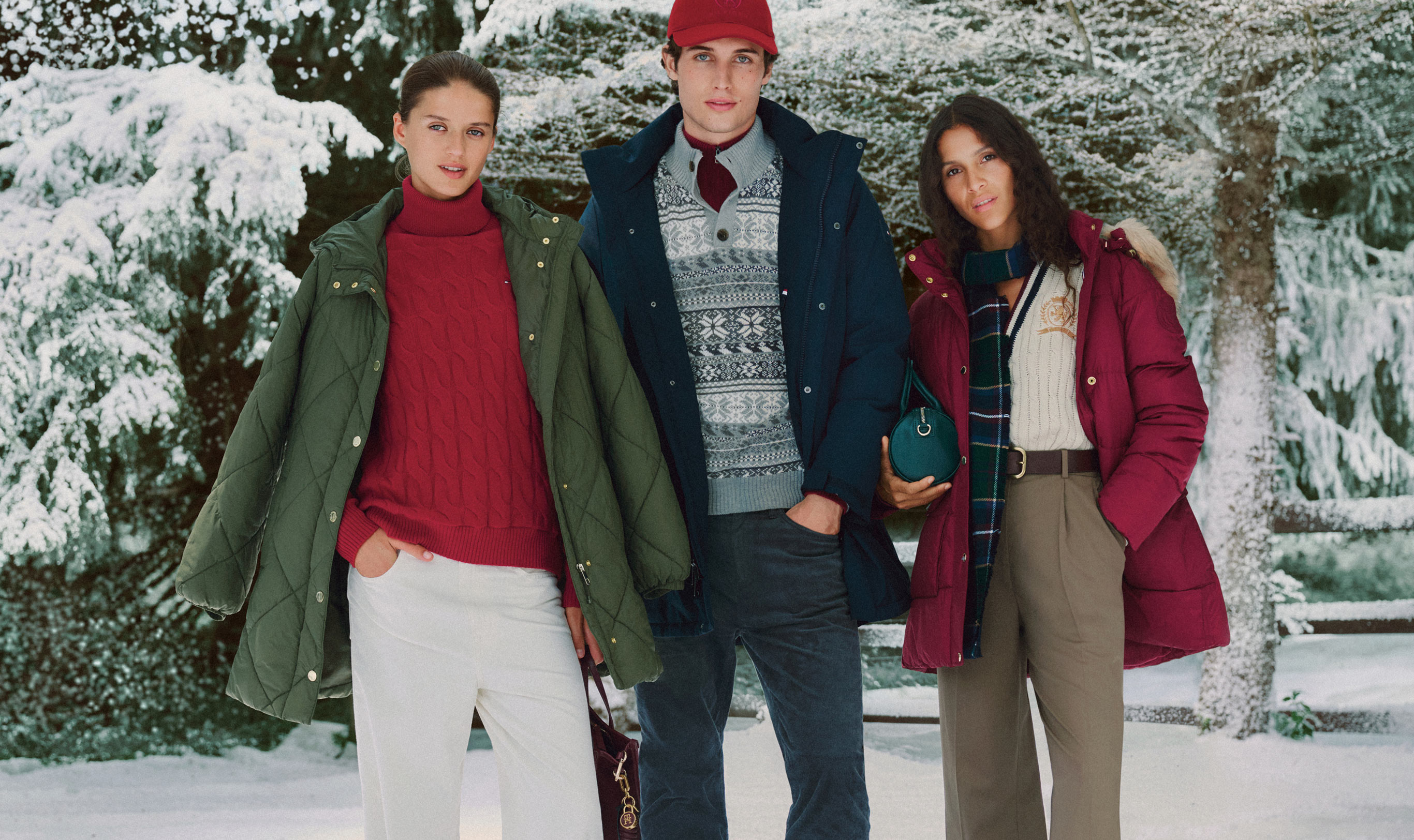 Three models walking past a snowy entryway in Tommy Hilfiger outerwear, including puffers, a parka, and layered knits with beanies.