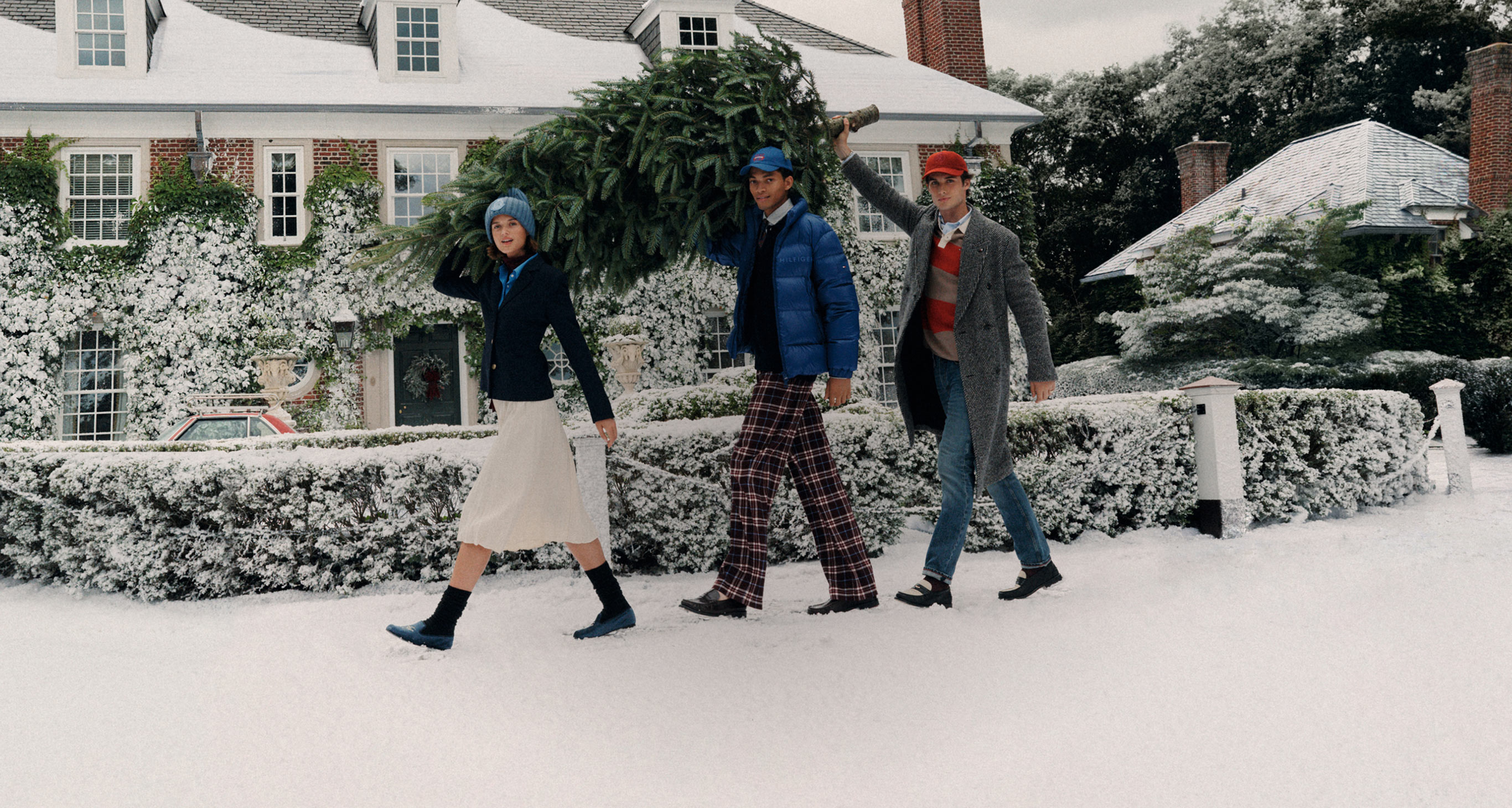 Group of models striding along a snow-covered xwsidewalk in Tommy Hilfiger coats and boots outside a festive house.