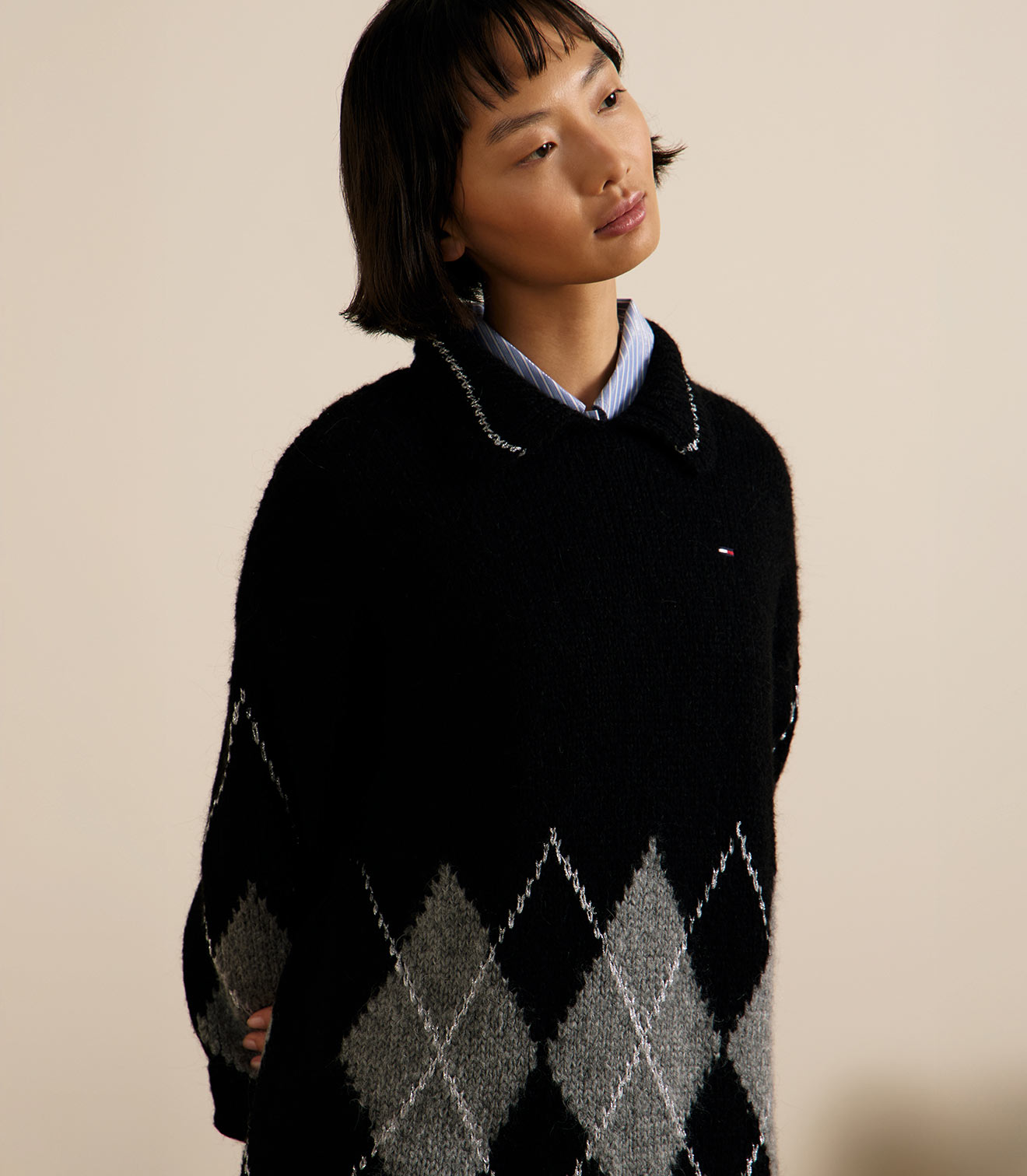 Model looking upward in a black Tommy Jeans sweater with gray argyle, layered over a collared shirt in a studio setting.