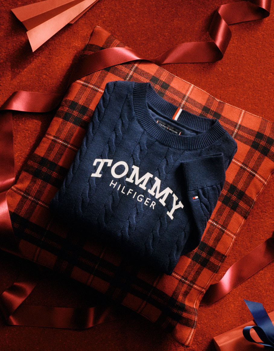 Folded Tommy Hilfiger logo sweater resting on a red plaid gift box with satin ribbons in a red studio setting.