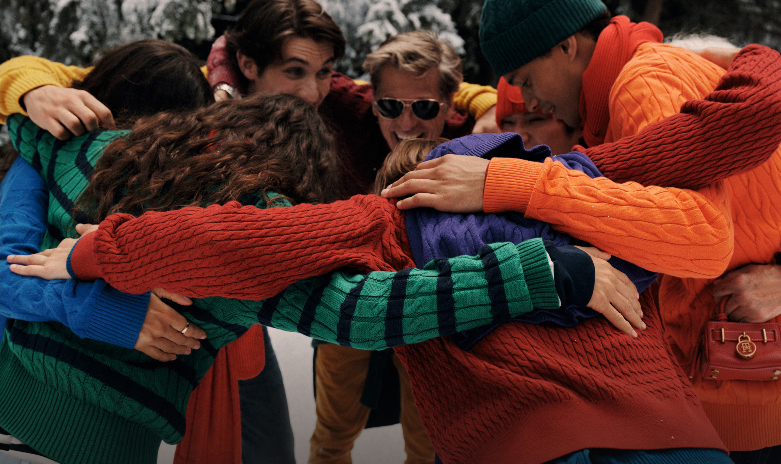 Close-up of friends in a group hug wearing Tommy Hilfiger cable-knit sweaters and scarves in bright orange, red, blue, and green. Text overlay reads 'Cables For All,' suggesting cozy winter layering.