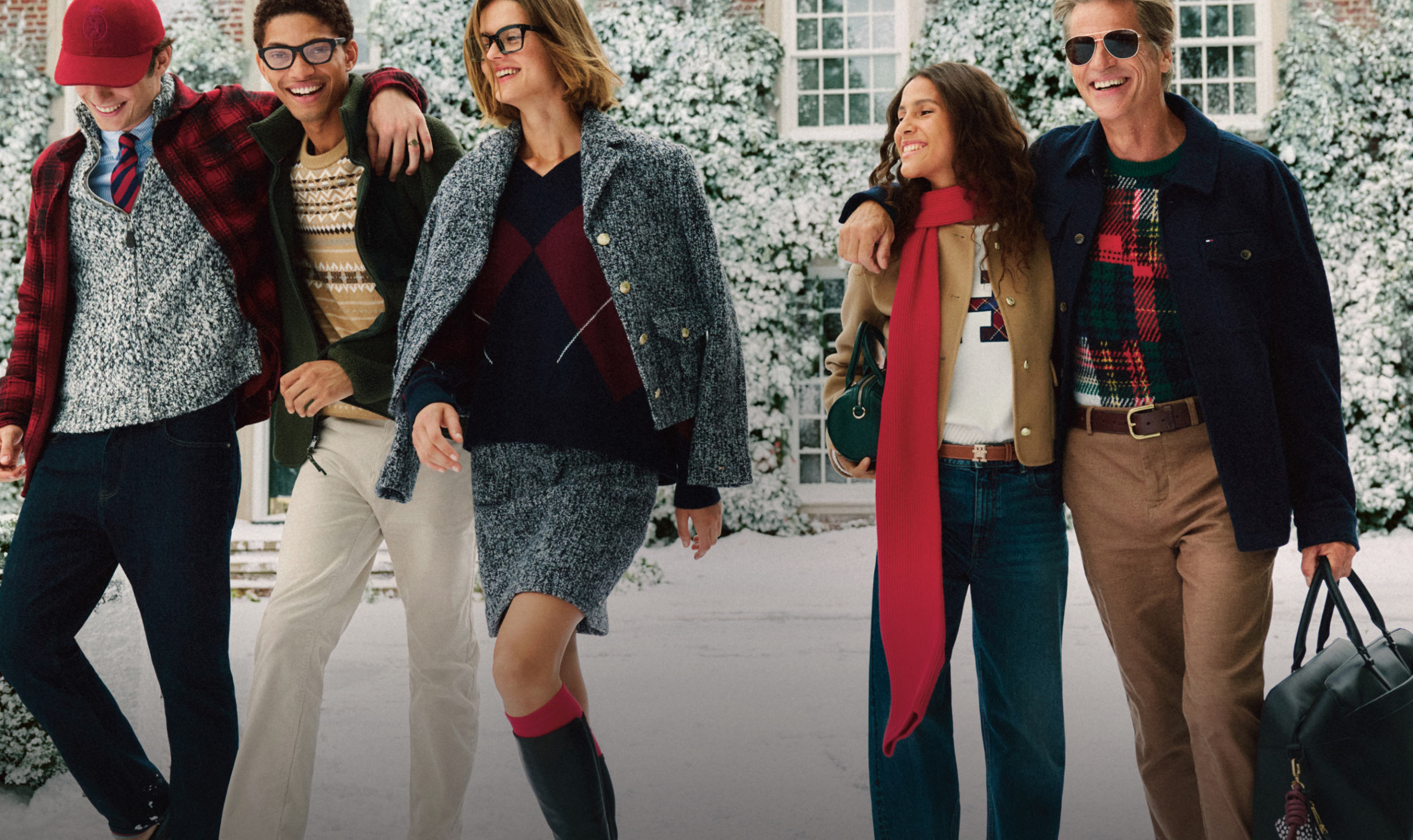 Group of models walking outside a snowy house wearing Tommy Hilfiger bottoms—dark wash jeans, tan chinos, and red trousers—styled with winter coats, patterned shirts, and scarves.