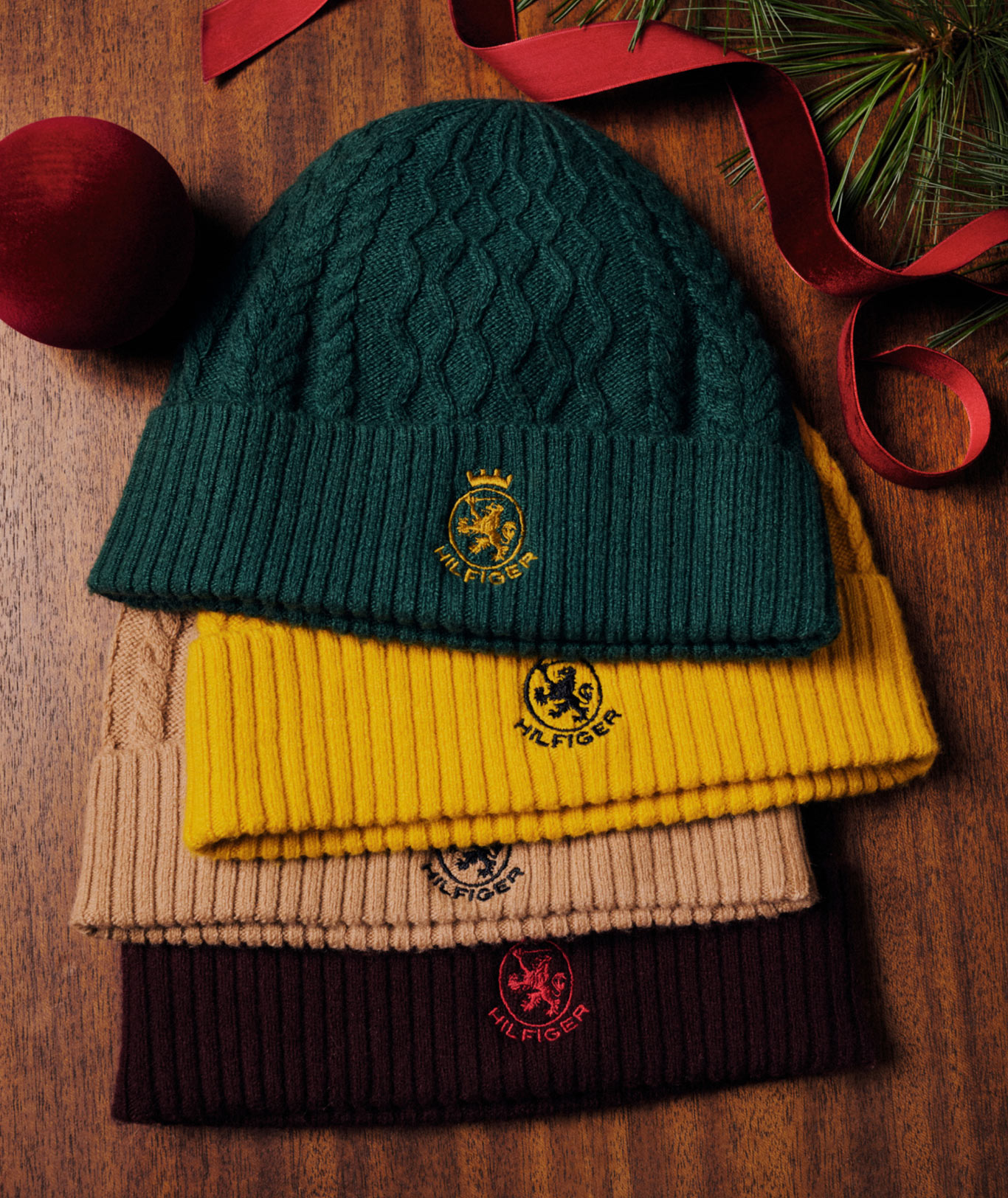 Stack of Tommy Hilfiger knit gifts on a wooden table, including crest beanies, folded sweaters, and scarves in green, mustard, pink, and red under holiday lights.