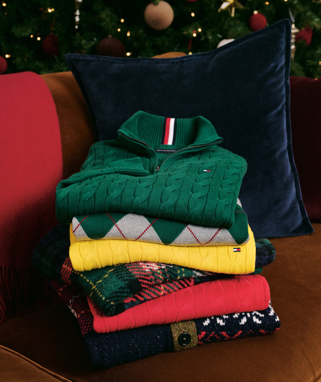Stack of Tommy Hilfiger knit gifts on a wooden table, including crest beanies, folded sweaters, and scarves in green, mustard, pink, and red under holiday lights.