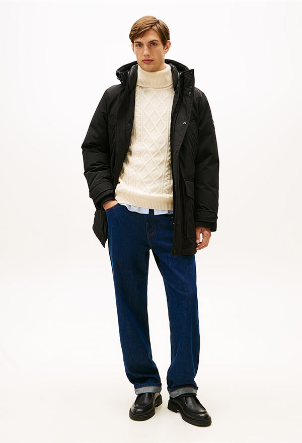 Model wearing a Tommy Hilfiger navy quilted puffer jacket layered over a yellow sweatshirt, styled with blue jeans and white sneakers, standing against a white background.