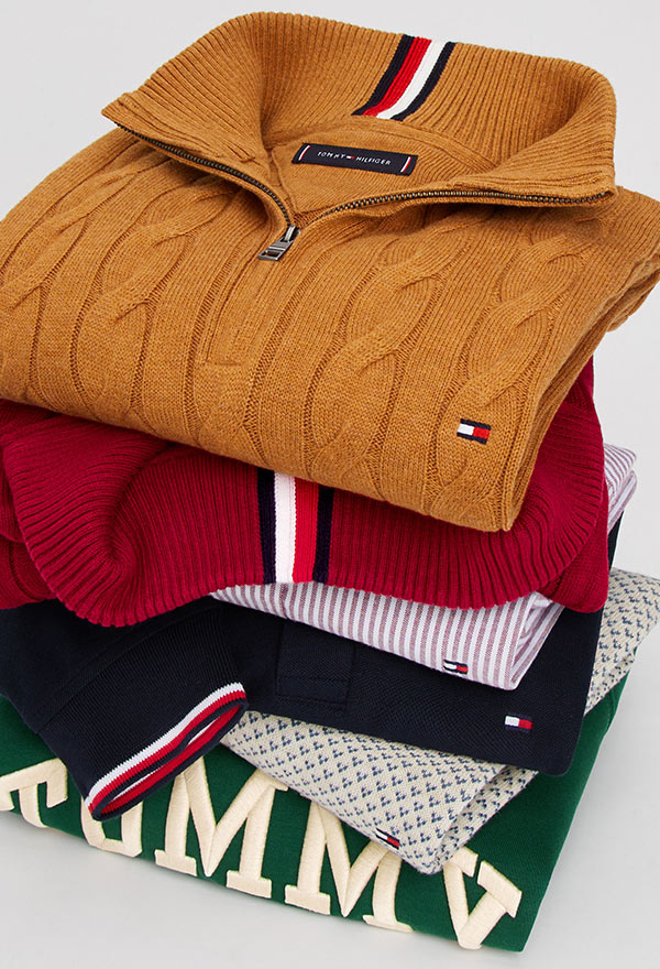 Stack of folded Tommy Hilfiger men's shirts, including an olive green polo, light blue button-down, navy logo tee, gray sweatshirt, and beige cable-knit sweater, showcasing signature flag branding