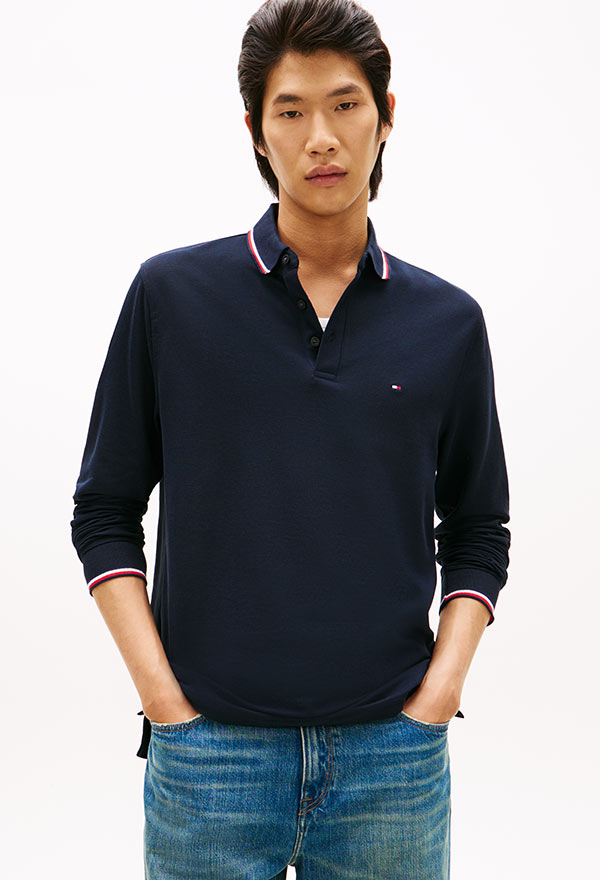 Man wearing a beige cable-knit sweater layered over a striped button-down shirt, paired with white pants, standing against a white background.