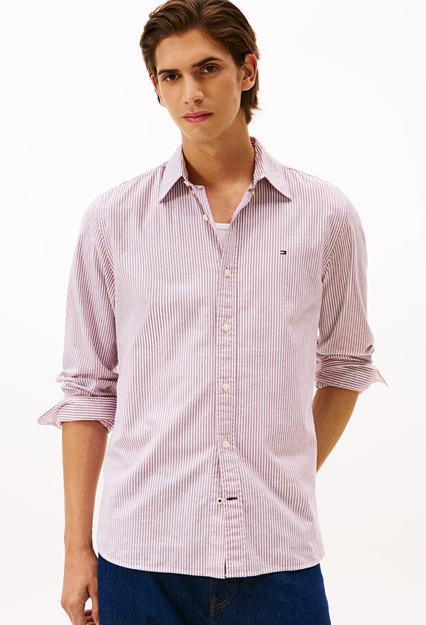 Model wearing a Tommy Hilfiger light blue long-sleeve button-down shirt with embroidered logo, paired with dark jeans, standing against a white background.