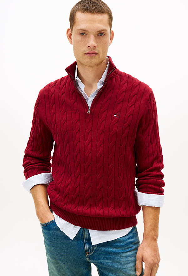 Man wearing a red cable-knit sweater layered over a striped button-down shirt, paired with white pants, standing against a white background.