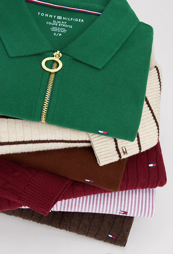 Stack of folded Tommy Hilfiger women's tops including a camel polo, blue sweater, grey knit, and navy piece, each featuring distinct textures and logo accents.