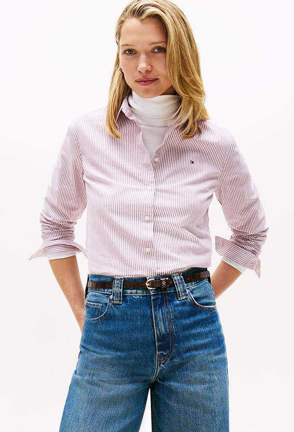 Model wearing a Tommy Hilfiger beige collared shirt with rolled sleeves, styled with a high-waisted denim skirt, posed against a white background.