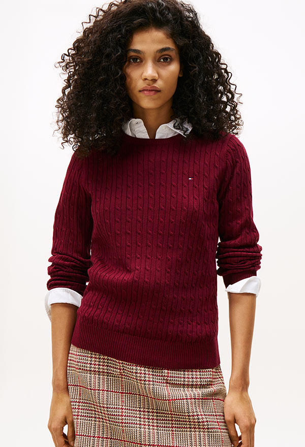 Model wearing a Tommy Hilfiger navy crewneck sweater with rolled sleeves and light blue jeans, standing against a white background.