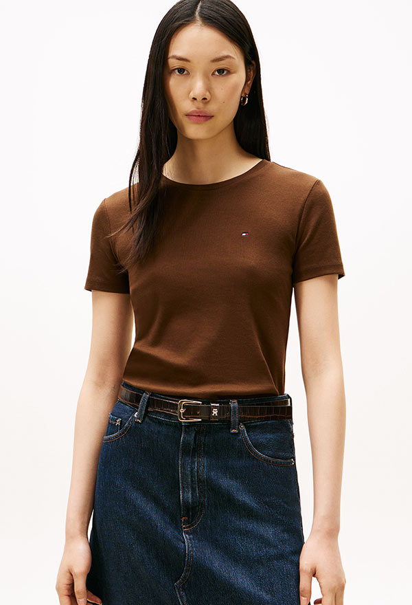 Model wearing a Tommy Hilfiger bright blue crewneck T-shirt tucked into high-rise white pants, standing against a white background.