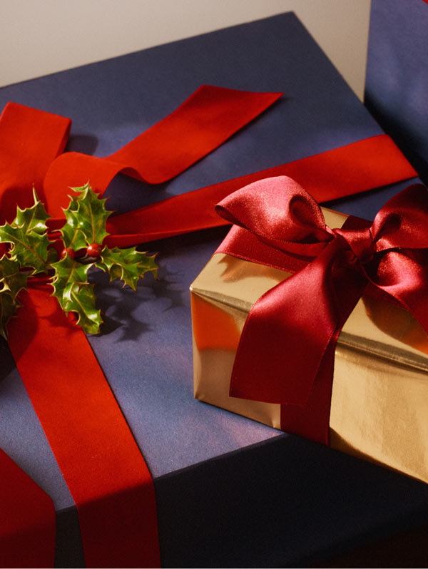 Close-up of a Tommy Hilfiger gift box wrapped in navy paper with a bold red ribbon and small greenery accent, styled as a festive holiday present.
