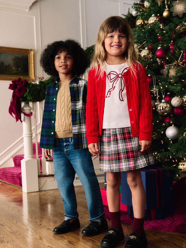Two children in festive Tommy Hilfiger holiday outfits standing in front of a decorated Christmas tree, wearing layered knits and plaid pieces.