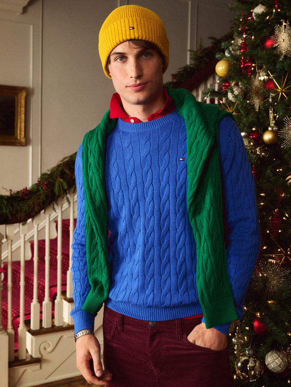 Model standing indoors by a decorated Christmas tree in a bright blue Tommy Hilfiger cable-knit sweater, with a green sweater draped over the shoulders and a yellow beanie.