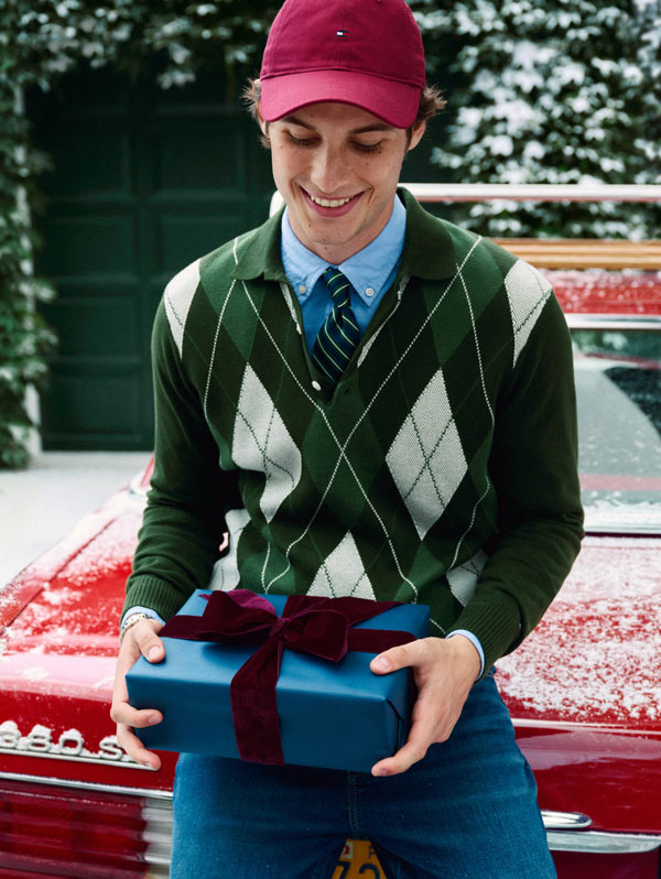 Model sitting on a car hood holding stacked gift boxes, wearing a green argyle Tommy Hilfiger sweater over a button-down shirt and a red baseball cap in a snowy outdoor setting.