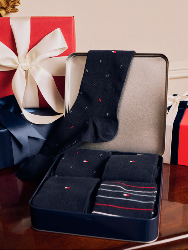 Open Tommy Hilfiger gift box with neatly arranged navy socks and accessories on a wooden surface, styled with large wrapped presents in the background.