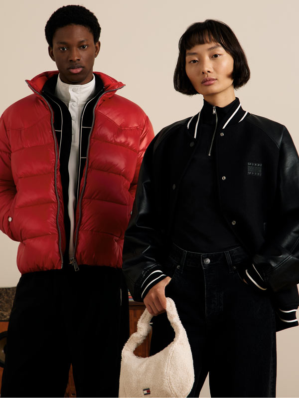 Two models in Tommy Hilfiger outerwear, one wearing a red puffer jacket and the other in a black jacket, standing side by side against a neutral studio backdrop.