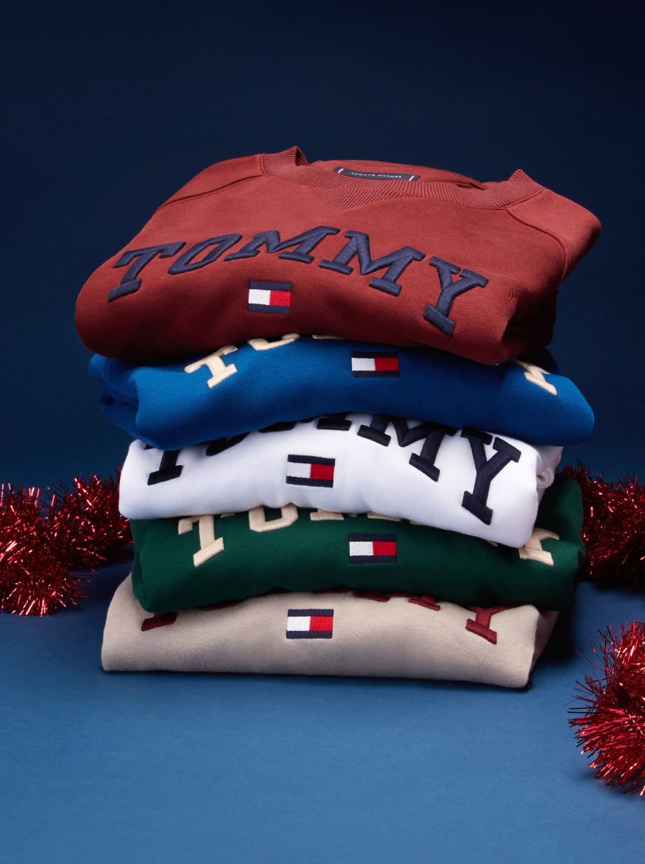Stack of folded Tommy Hilfiger holiday sweaters with bold logo and festive motifs, styled on a blue surface with red tinsel garland.