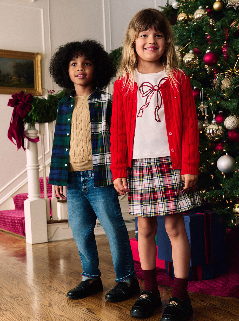 Two children in festive Tommy Hilfiger holiday outfits standing in front of a decorated Christmas tree, wearing layered knits and plaid pieces.