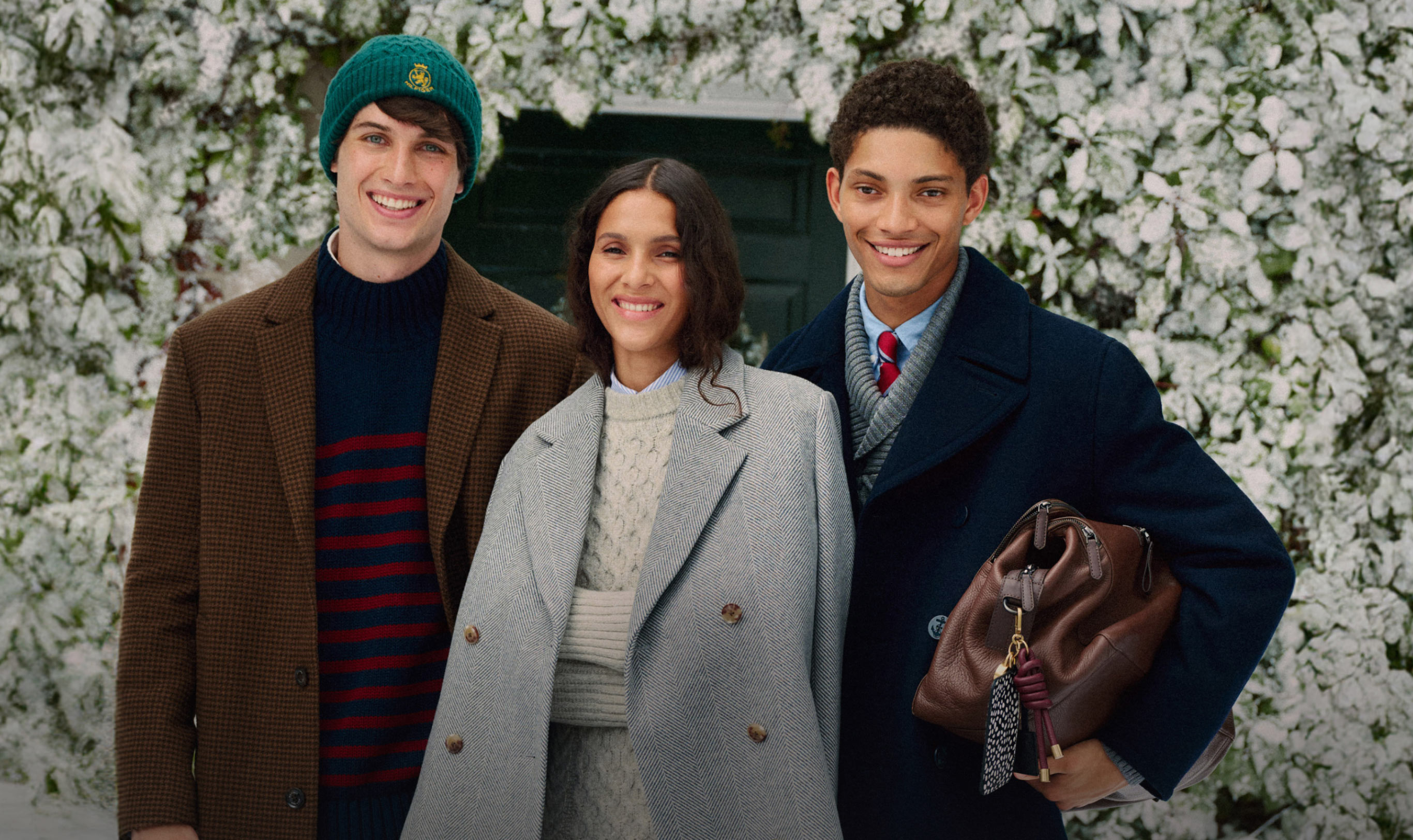 Three models in tailored Tommy Hilfiger winter coats and knit hats standing in front of a snowy, floral backdrop, smiling at the camera.