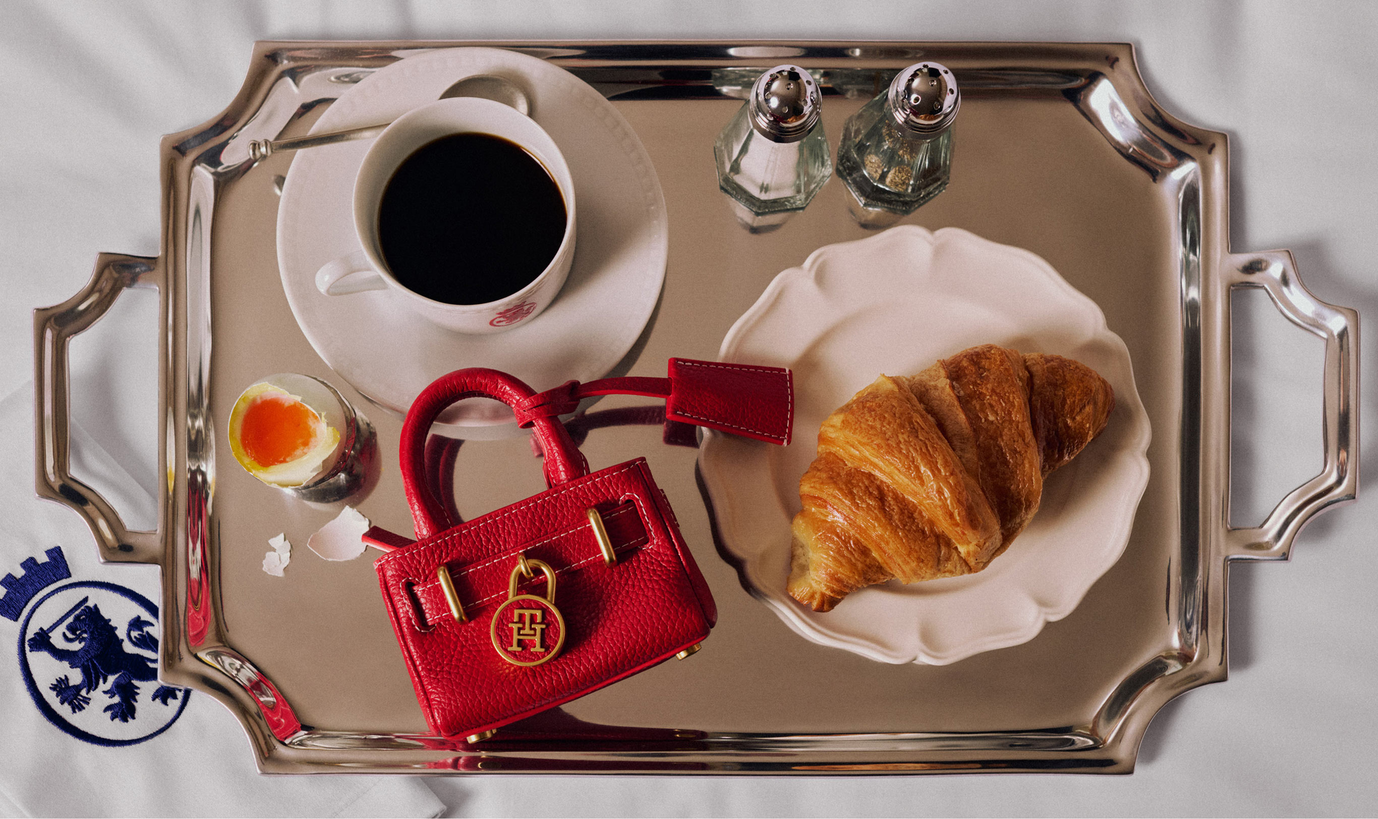 Overhead view of a silver tray with coffee, a pastry, and small accessories arranged beside a folded Tommy Hilfiger item, styled as a holiday gift moment.