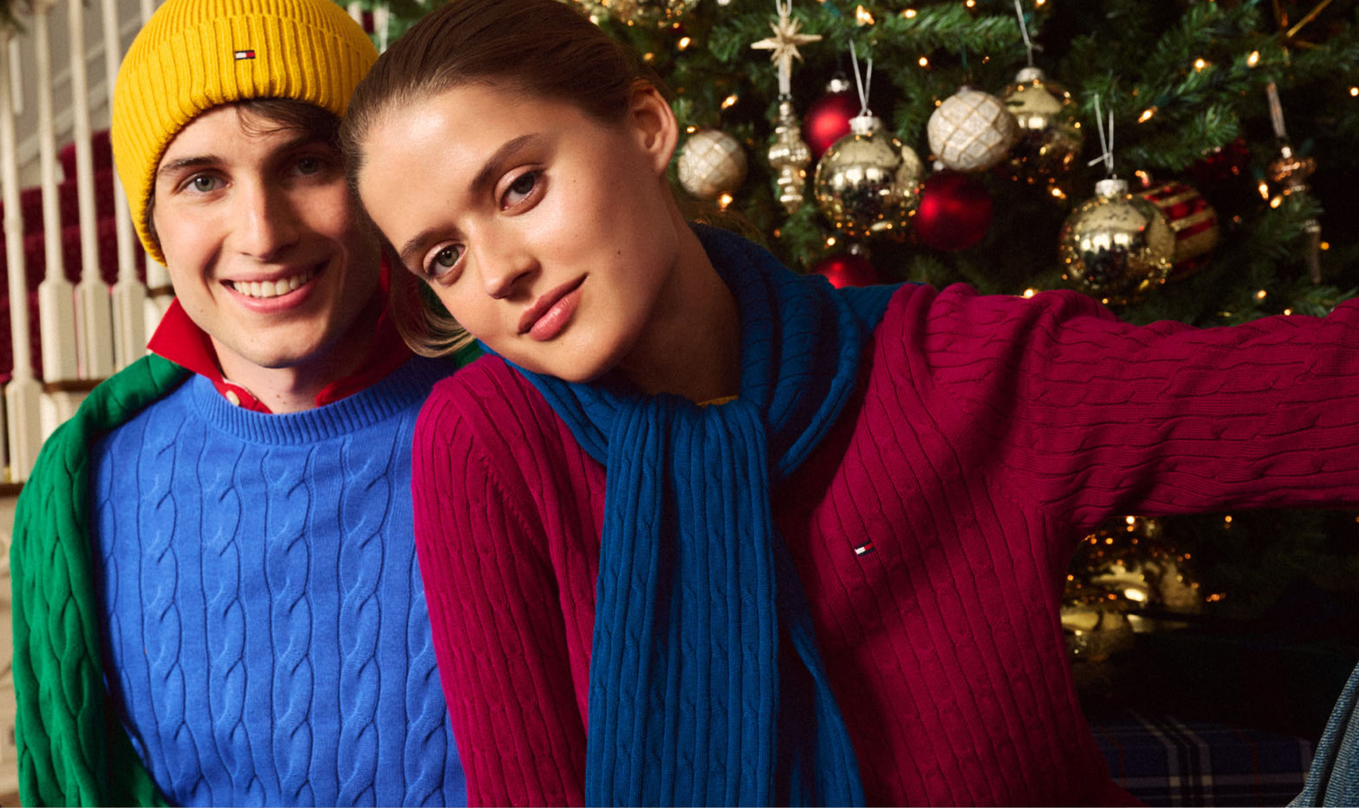Group of models in colorful Tommy Hilfiger holiday outfits, including bold knits and tailored layers, standing together and smiling in front of a decorated Christmas tree.