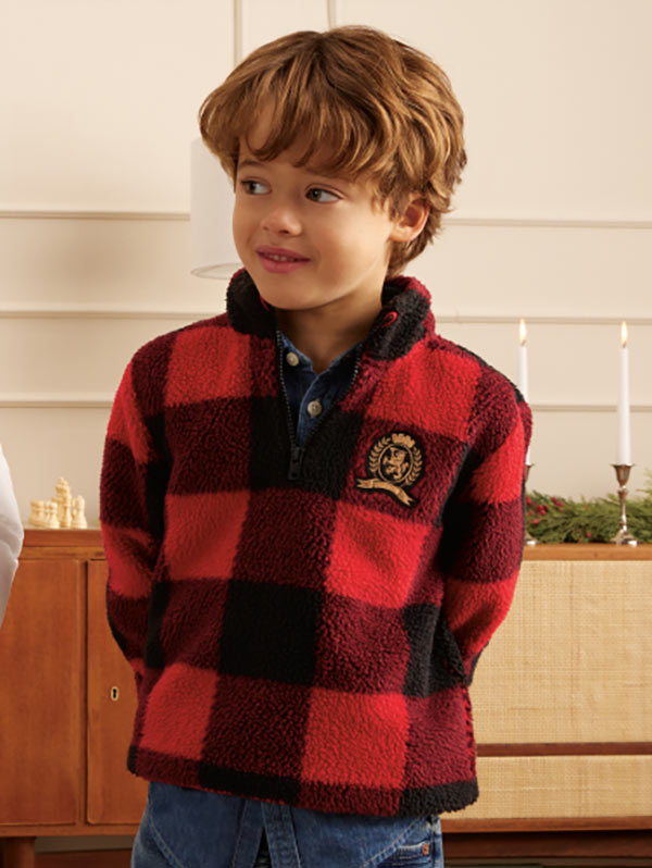 Young boy wearing a red and black check Tommy Hilfiger zip-up sweater with a crest logo, standing indoors by a wooden counter.