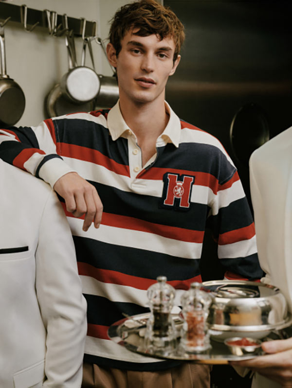 Male model leaning on a café counter wearing a striped rugby-style Tommy Hilfiger shirt with a bold crest logo, surrounded by coffee cups and metal canisters in a busy indoor setting.