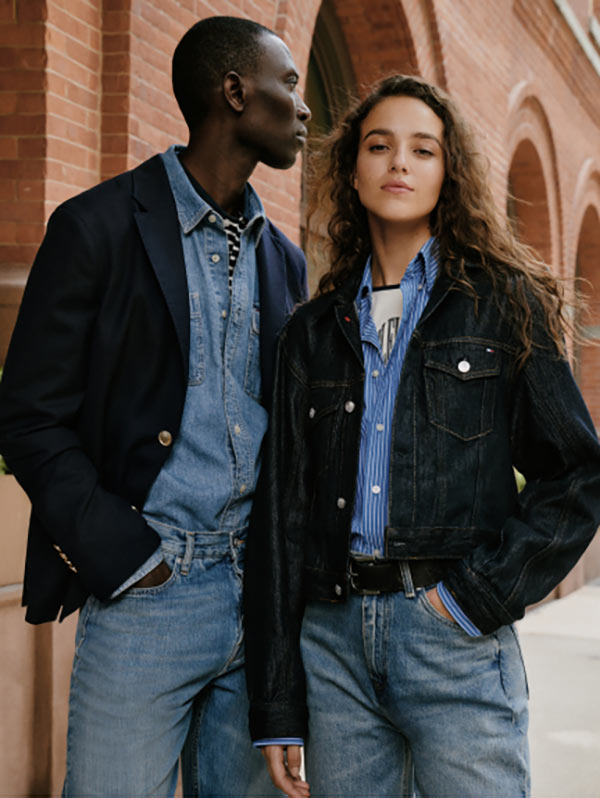 Two models standing outdoors against a brick building, wearing layered denim looks with a navy jacket and blue jeans on the left and a black denim Tommy Hilfiger jacket with light-wash Tommy Hilfiger jeans on the right.