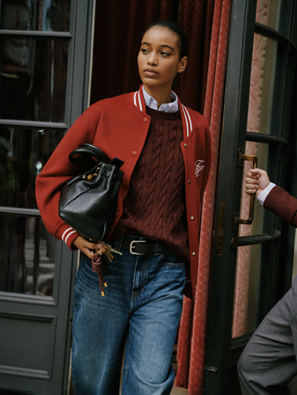 Female model stepping out of a doorway in a red Tommy Hilfiger jacket over a textured knit top and blue jeans, carrying a black Tommy Hilfiger handbag on a city street.