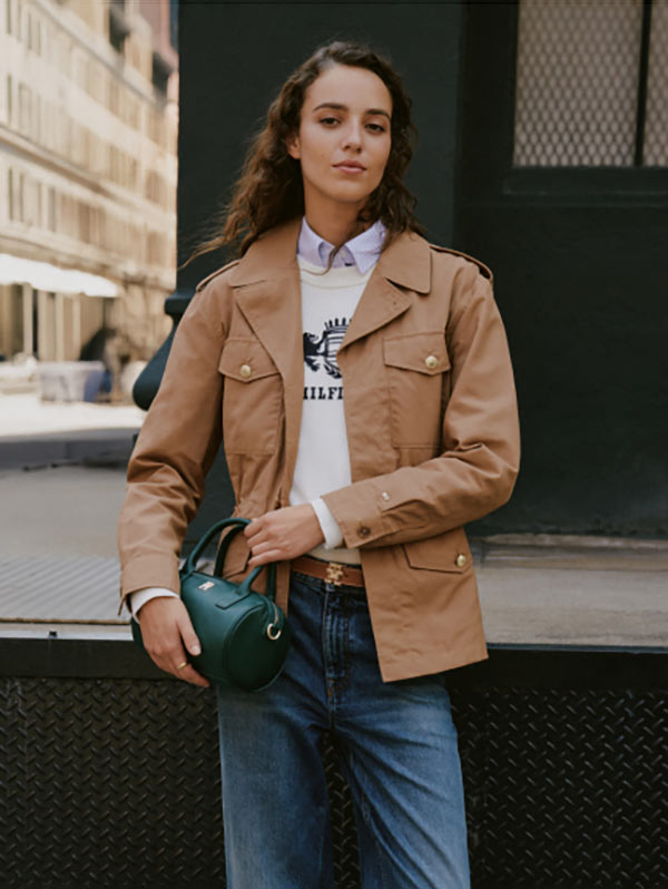Female model outdoors wearing a tan utility-style Tommy Hilfiger jacket over a white top with blue Tommy Hilfiger jeans, holding sunglasses and a small accessory while standing near a modern building facade.