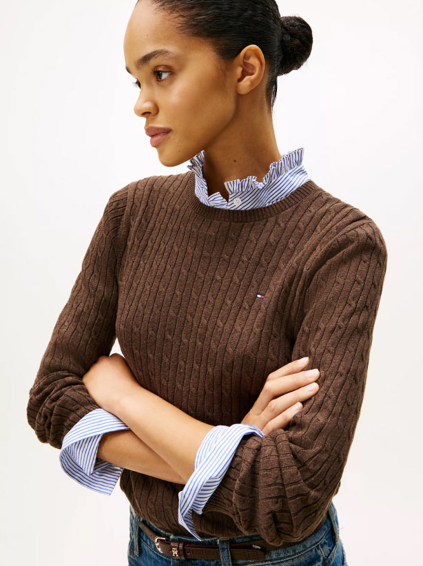 A model wearing a brown textured Tommy Hilfiger sweater layered over a light blue ruffled-collar shirt, with sleeves rolled and arms crossed.