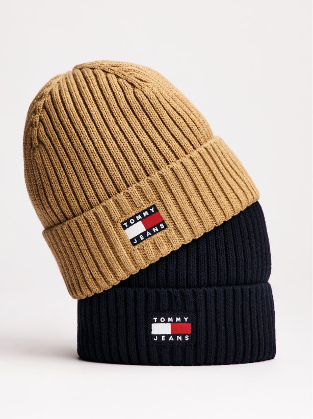 Two stacked Tommy Hilfiger beanies, with a camel ribbed knit beanie resting over a navy cuffed beanie, both featuring the signature Tommy Hilfiger flag logo on the front against a light background.