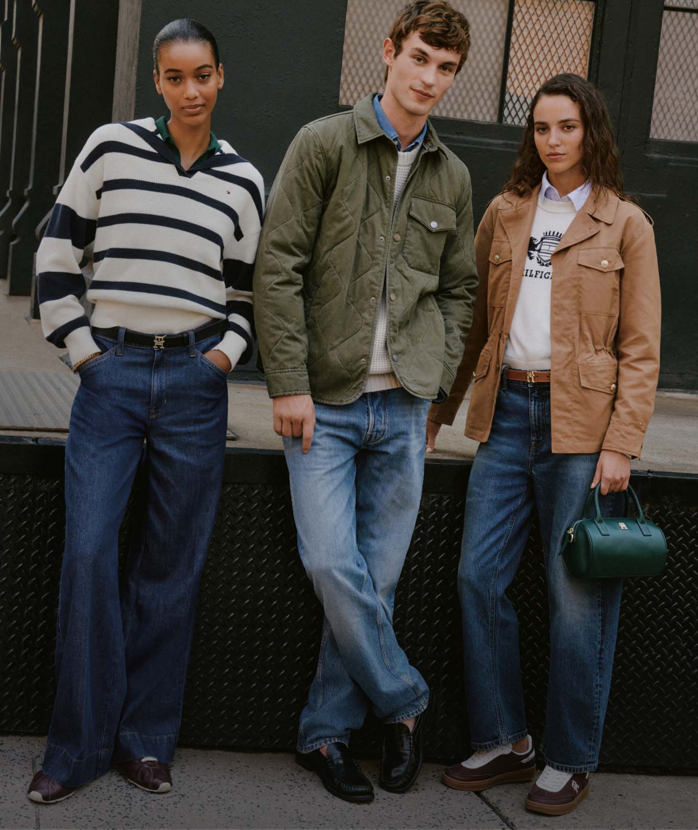 A group of models wearing Tommy Hilfiger striped sweaters, jeans, and jackets, standing together on a city street in a casual, collegiate look.