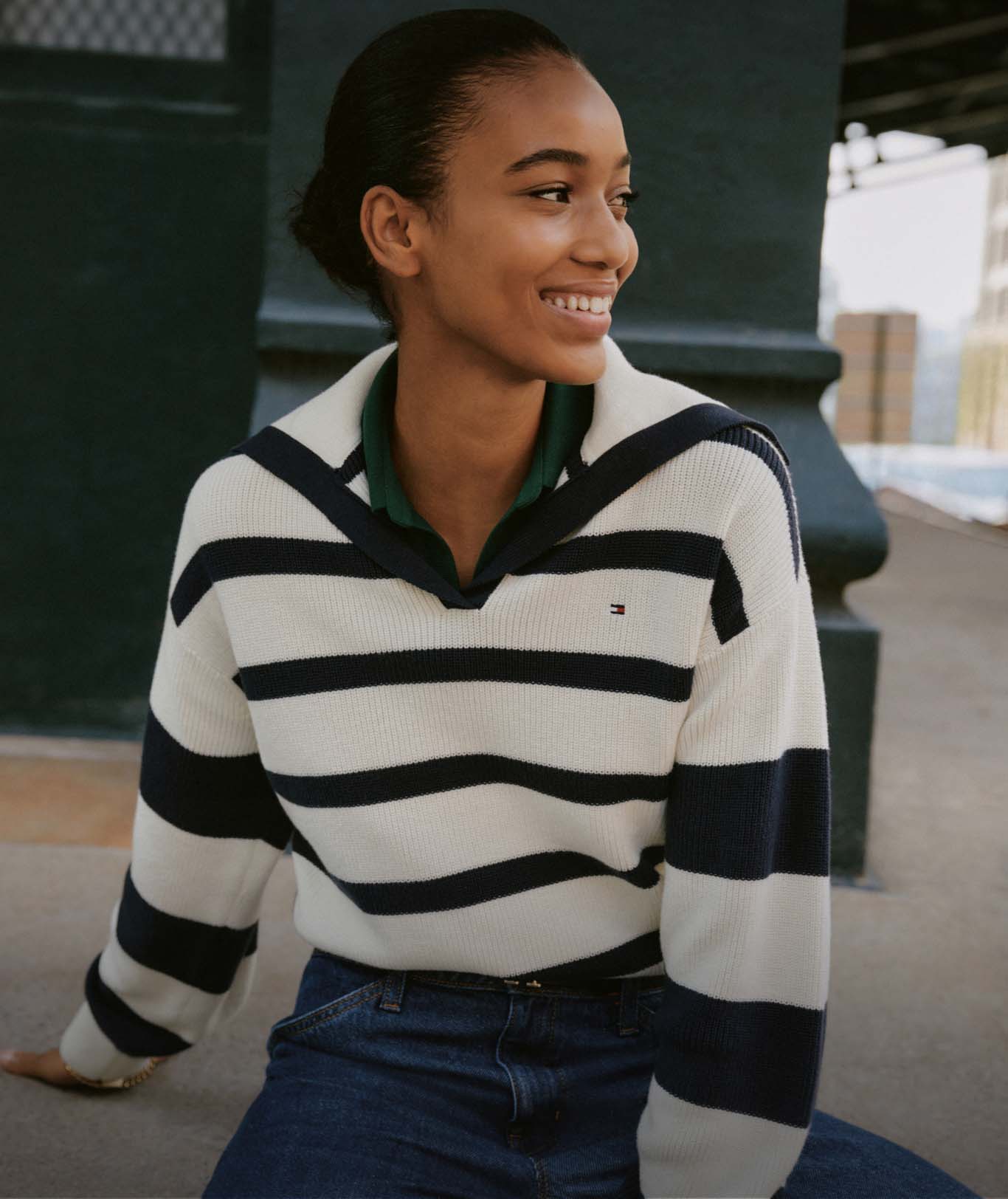 A group of models wearing Tommy Hilfiger striped sweaters, jeans, and jackets, standing together on a city street in a casual, collegiate look.