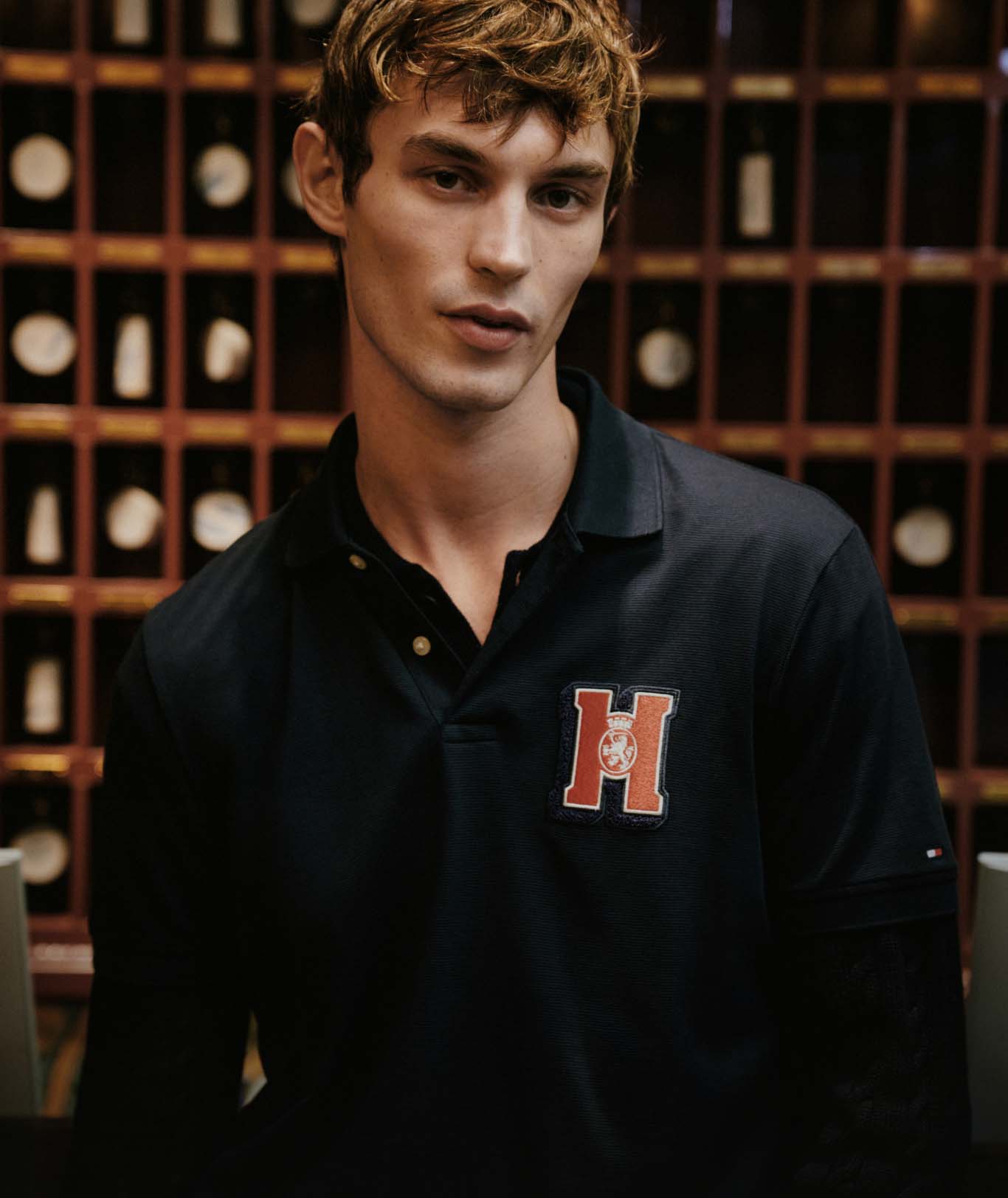 a male model in a navy polo with a varsity “H” logo, both wearing Tommy Hilfiger in a warm, indoor setting.