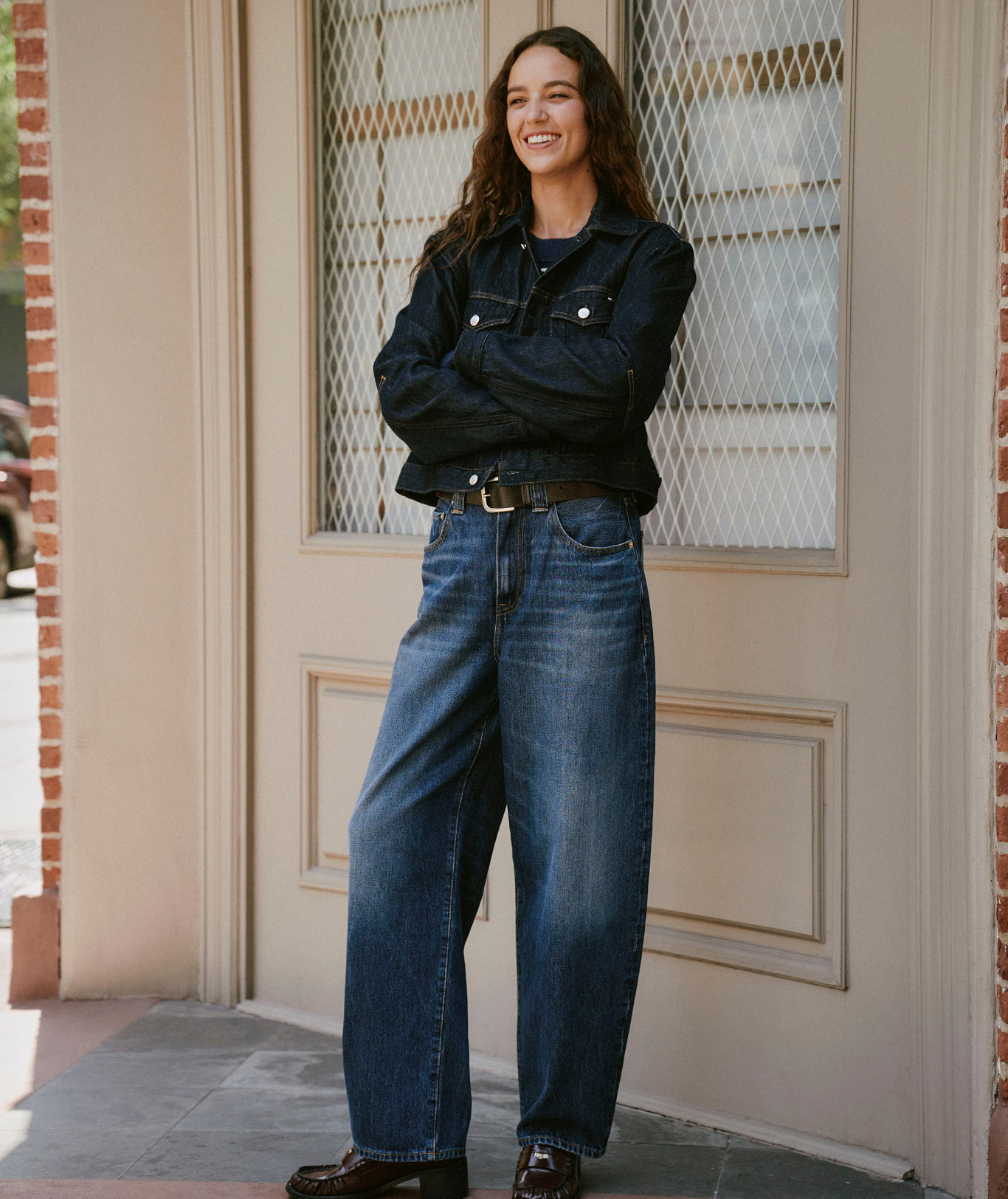 A model in Tommy Hilfiger denim and outerwear on a city street, featuring a dark jacket and wide leg jeans.