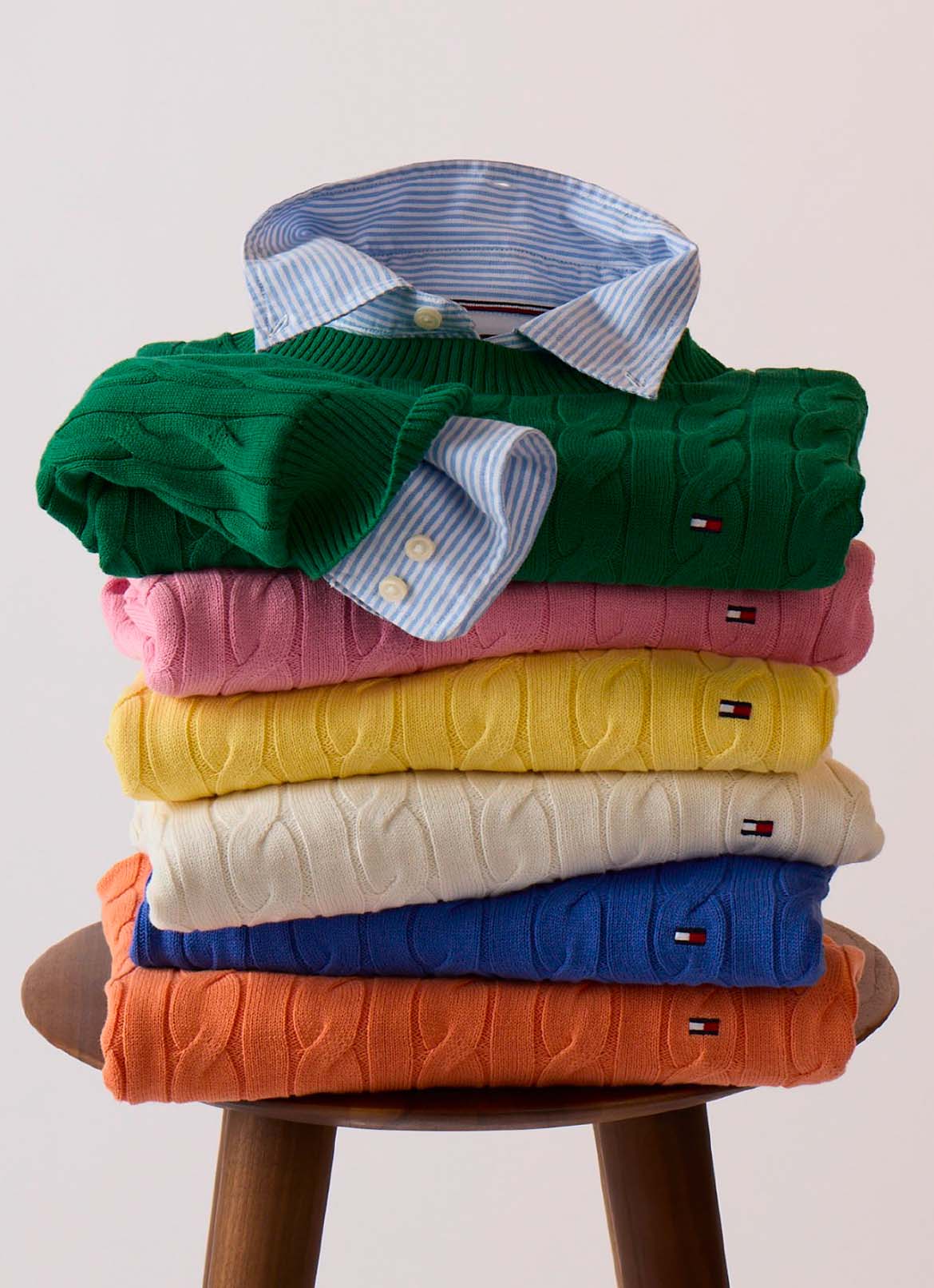 A stool stacked with folded Tommy Hilfiger knit sweaters in green, yellow, pink, and neutral tones in a studio setting.